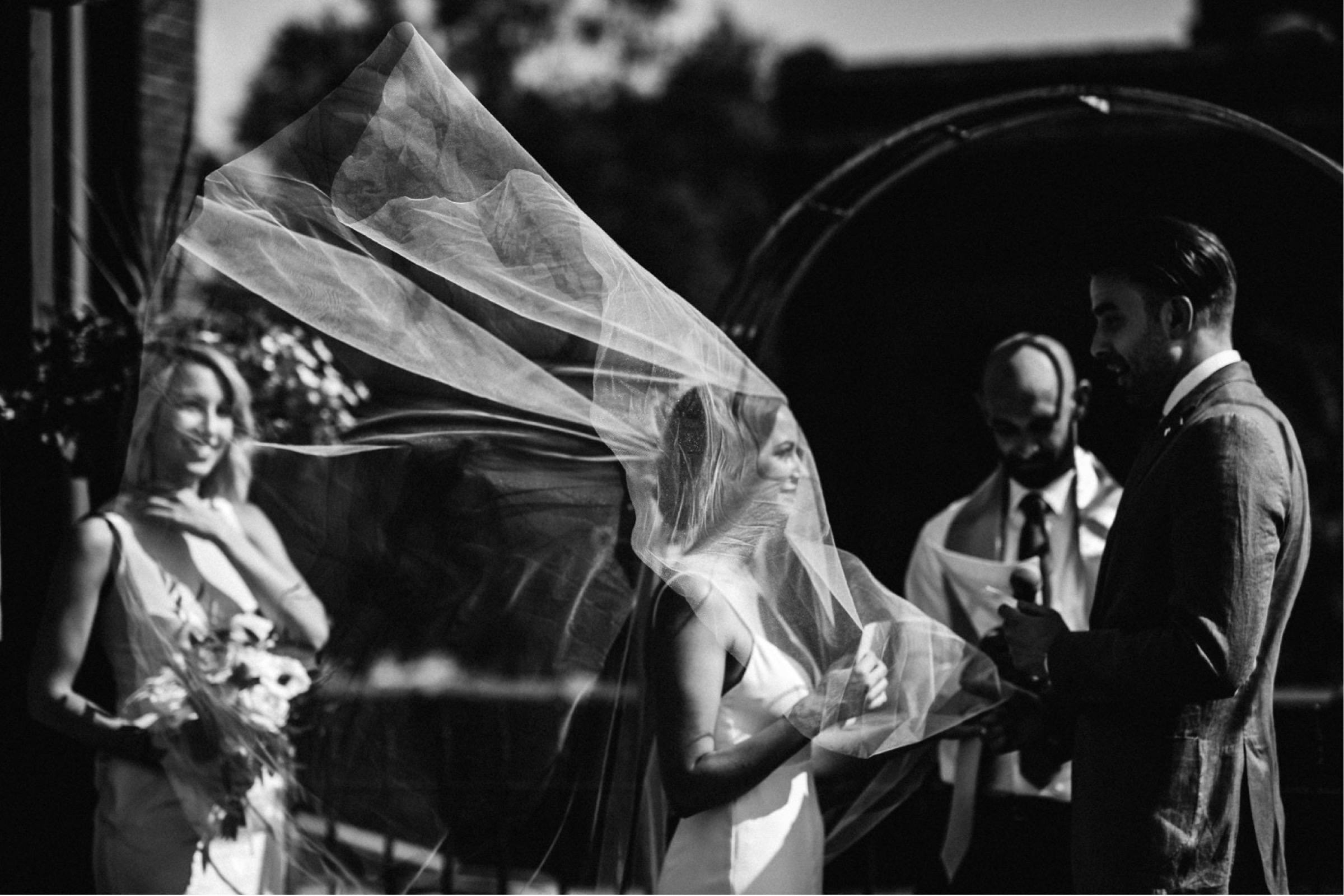 The wind is dramatically blowing a bride's veil behind her during a ceremony portion of a wedding day timeline