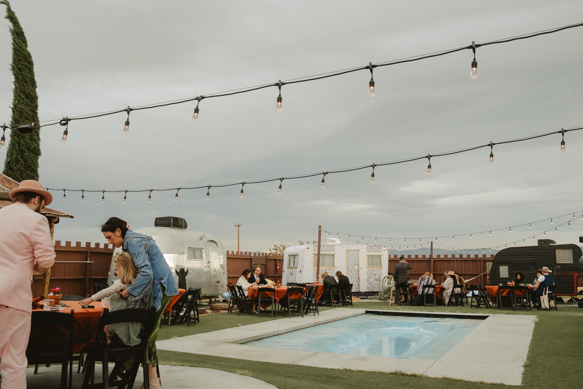 Outdoor gathering by a small pool, with people seated at tables. String lights are overhead, and vintage trailers are in the background. A person in a pink outfit is standing on the left.