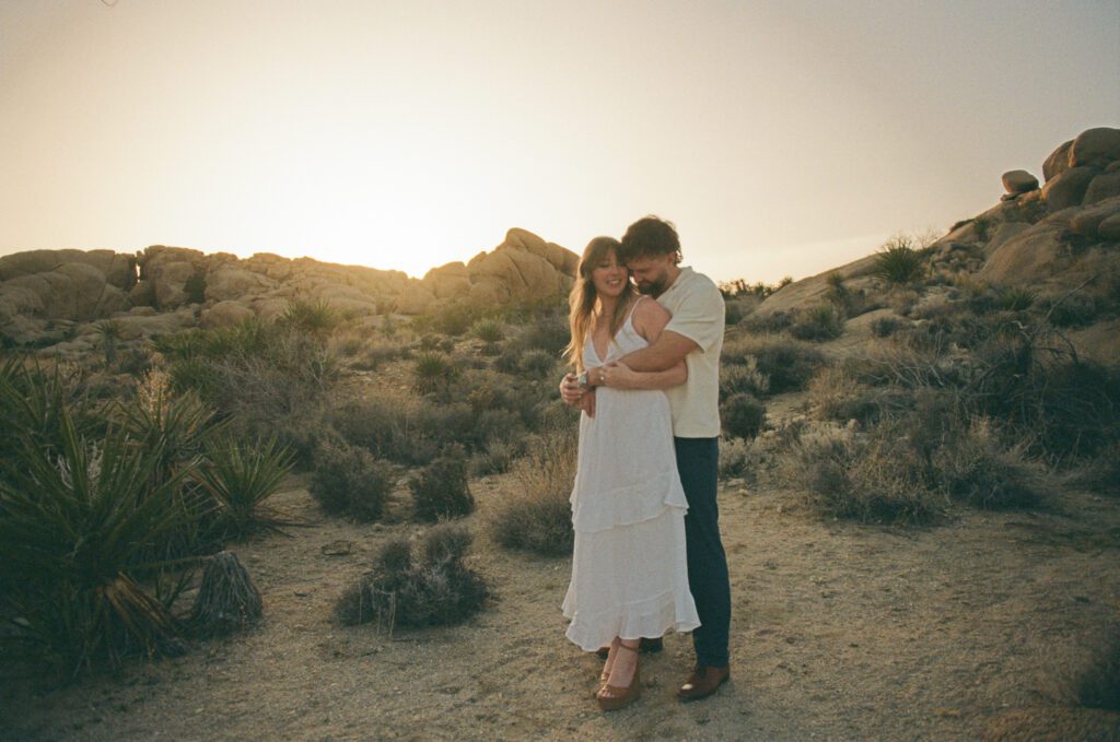 A couple embraces in a desert landscape at sunset, surrounded by rocks and sparse vegetation.