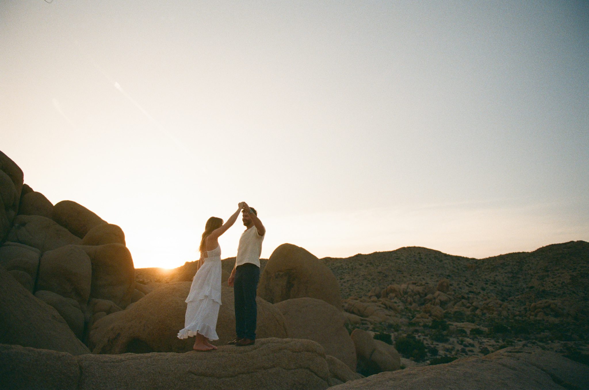 A couple dances on rocky terrain during sunset, with a clear sky and distant hills in the background.