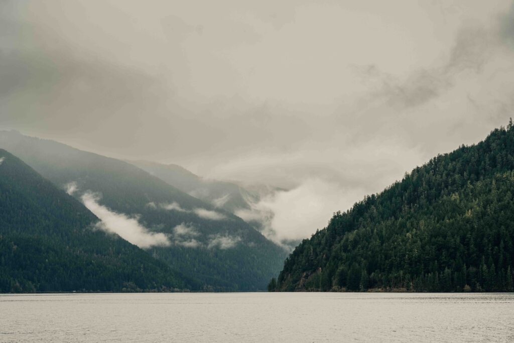 Cloudy sky over a serene lake surrounded by forested hills and misty mountains.