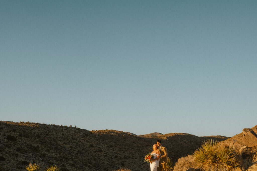 A couple stands in a desert landscape at sunset, holding a bouquet. The sky is clear, and the surroundings are rocky with sparse vegetation.