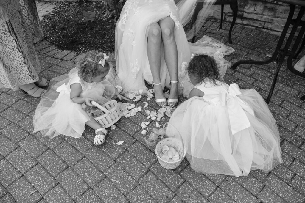 Two young girls in dresses gather petals from fallen flower baskets, while a seated woman in a gown looks on, outside on a paved area.