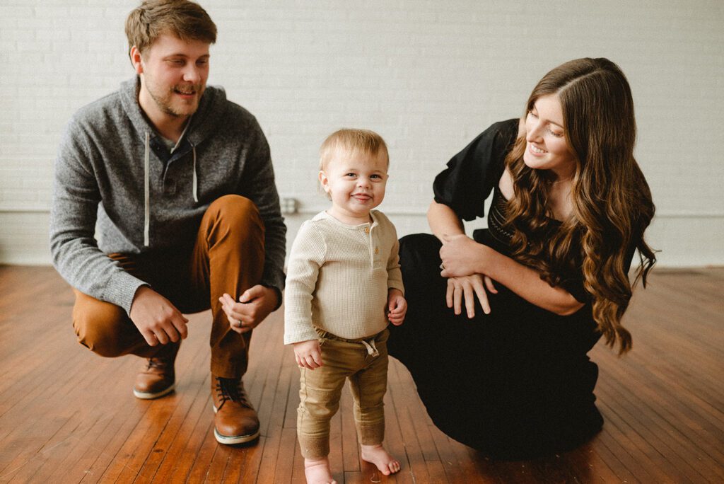 A toddler stands smiling between two adults, one kneeling and one crouching, on a wooden floor with a white brick wall backdrop.