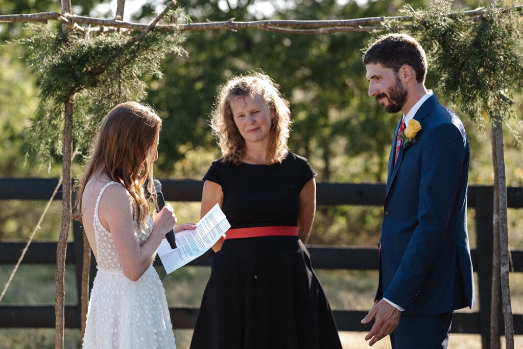 A woman in a white dress reads from paper, standing beside a person in a black dress and a man in a suit with a yellow flower, in an outdoor setting with wooden arch and greenery.