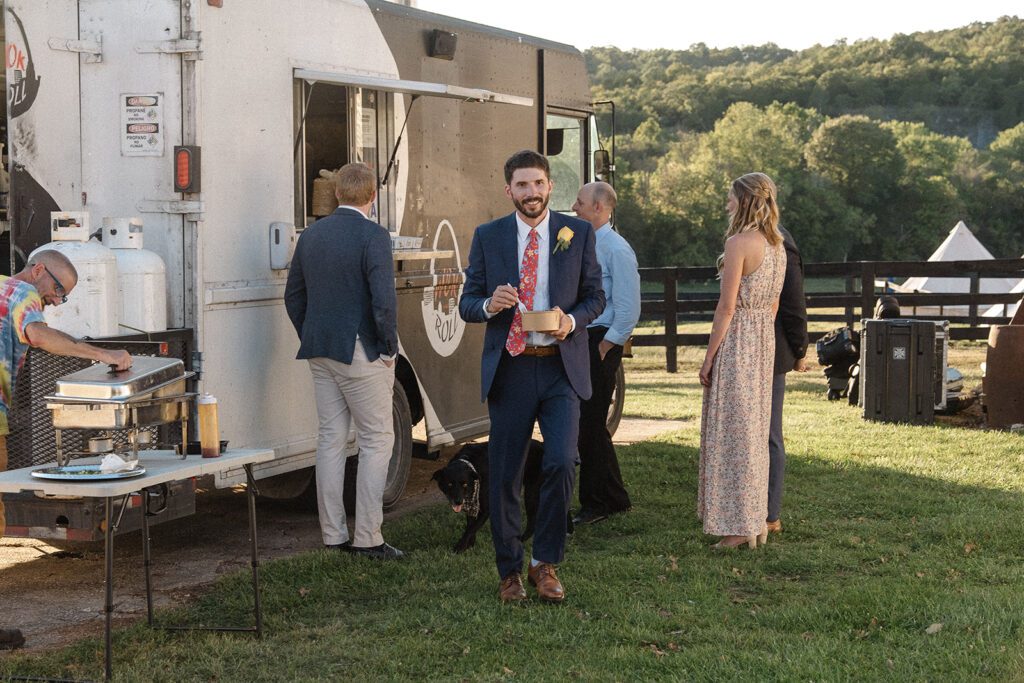 A man in a suit holds a plate near a food truck at an outdoor event. Others stand nearby, including a person serving food. Trees and grass are in the background.