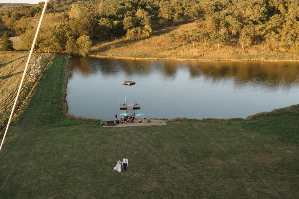 A couple walks hand in hand across a large grassy field toward a small dock on a calm lake. Trees line the background under a clear sky.