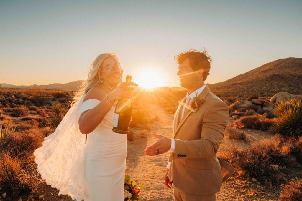 A bride and groom in wedding attire stand in a desert landscape at sunset. The bride is holding a bottle of champagne and the groom is extending his hand.