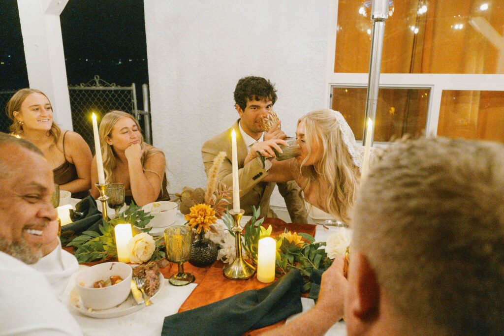 A smiling bride and groom are seated at a table with six guests during their destination wedding in Joshua Tree. The bride playfully feeds the groom a drink. The table is decorated with candles and flowers.