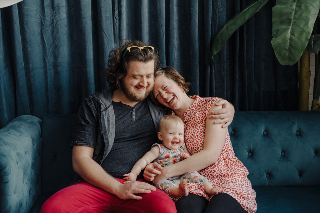 A couple sits on a blue couch with a smiling baby between them, embracing and smiling.