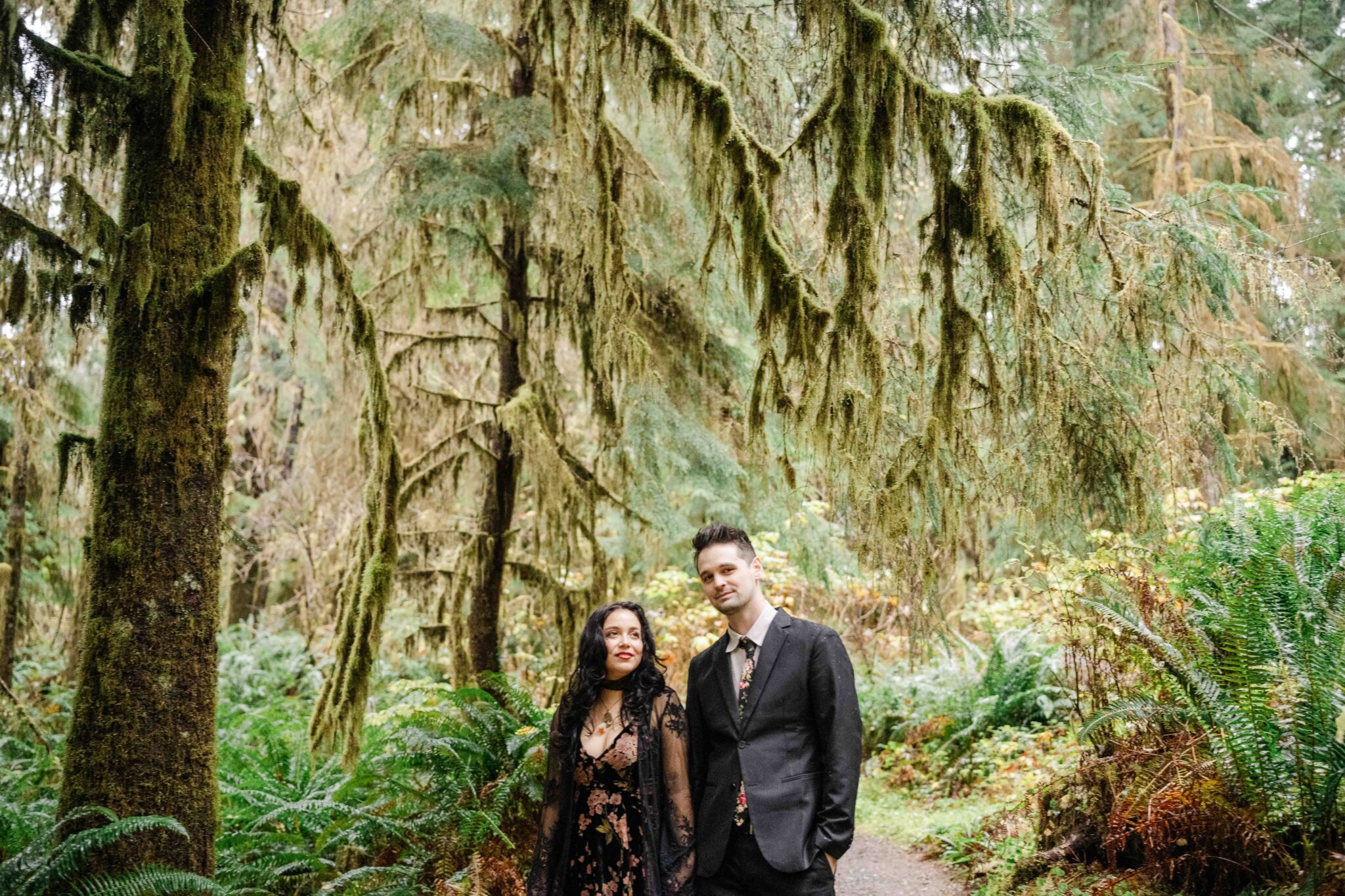 A man and a woman standing on a forest path surrounded by lush trees and greenery, with the man wearing a suit and the woman in a patterned dress.