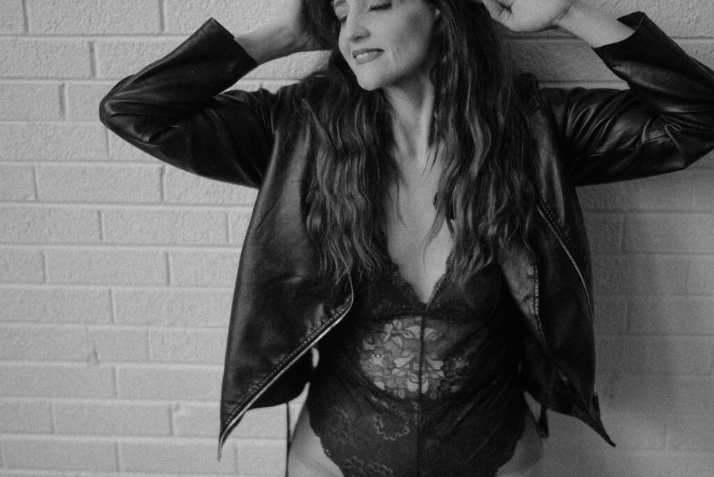 A woman in a black lace outfit and leather jacket stands against a white brick wall, with eyes closed and hands touching her head.