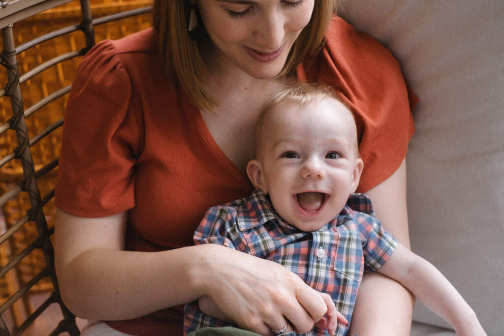 A woman in an orange shirt holds a smiling baby wearing a plaid shirt while sitting on a couch.