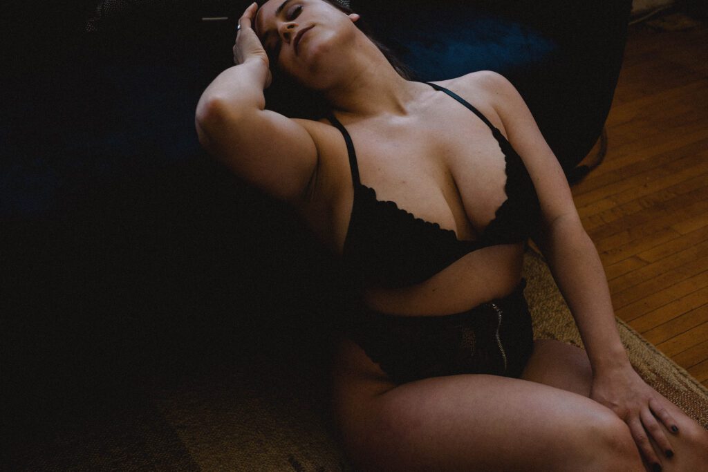 Person sitting on the floor in dark lighting, wearing a black bra and high-waisted bottoms, with head tilted back and one hand touching their head.