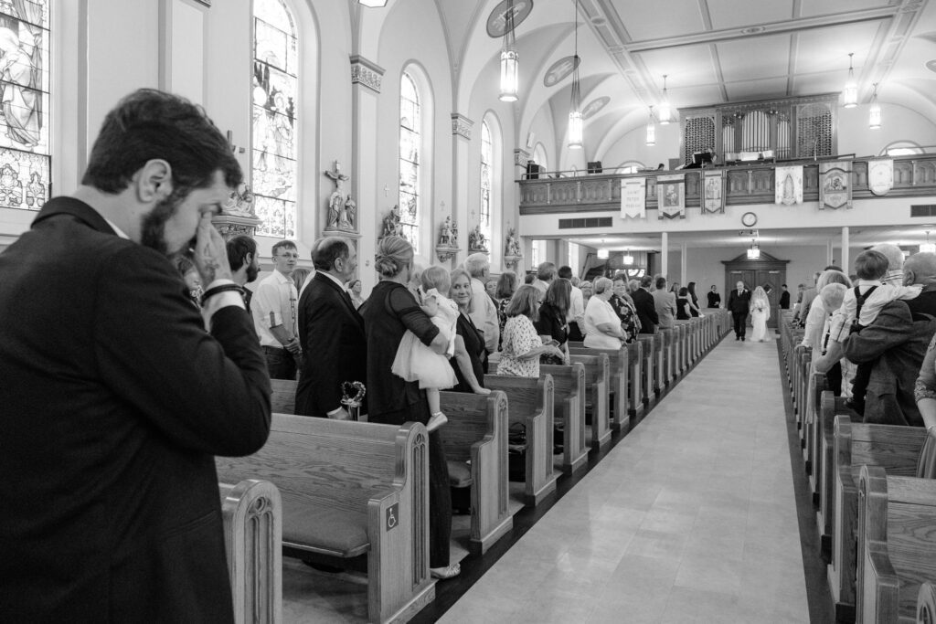 A person wipes their eyes in the foreground while a wedding ceremony proceeds in a church, with guests seated in pews and a couple standing at the altar.