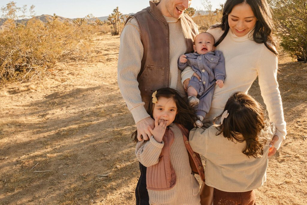 A family of four stands outdoors in a desert landscape; two young girls, a baby held by the mother, and an adult man, all dressed in warm clothing.