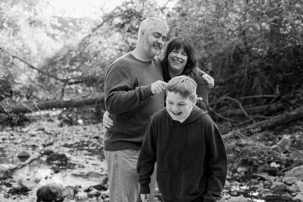 A man, woman, and boy stand together outdoors near a creek, smiling and laughing among trees and rocks.