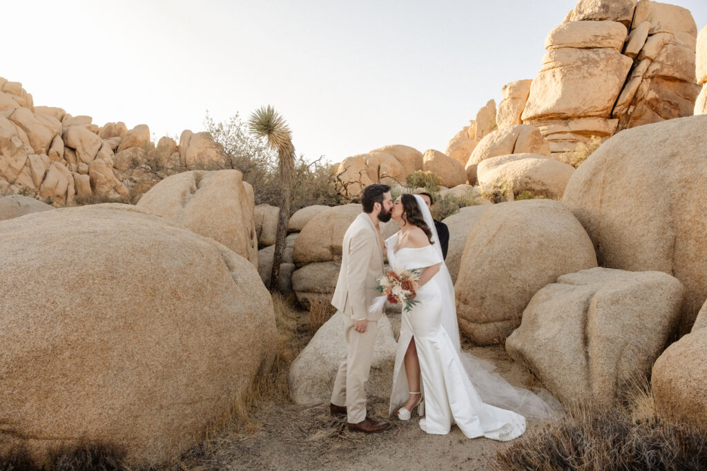 A bride and groom kiss during their Joshua Tree Micro Elopement, standing among large desert rock formations, dressed in wedding attire and holding a bouquet.