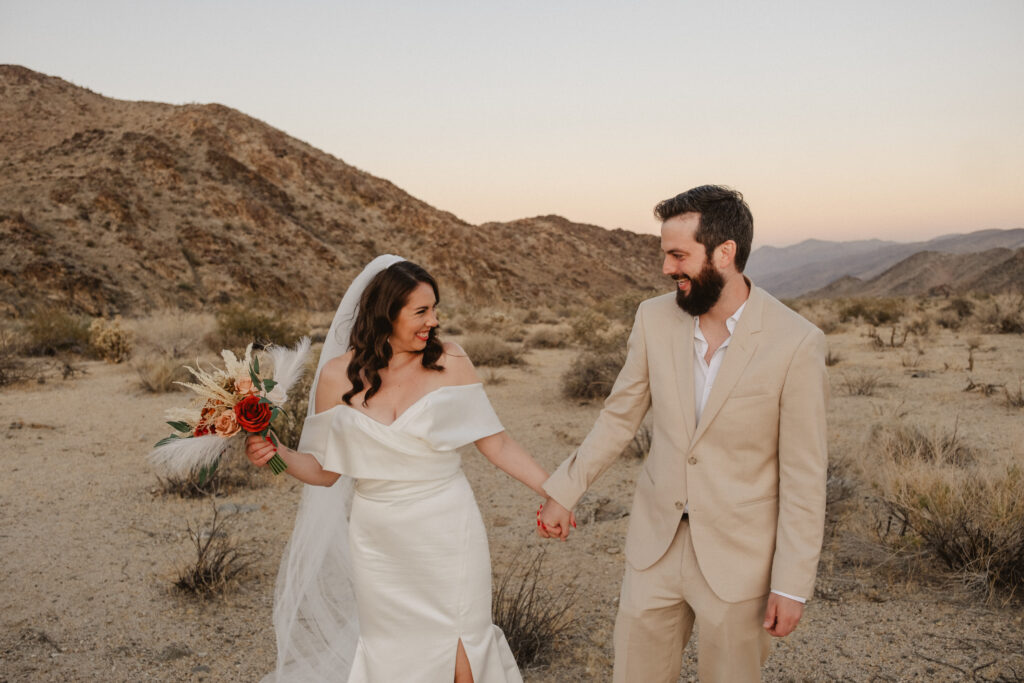 A bride in a white gown and veil holds a bouquet and the hand of a groom in a beige suit as they stand in a desert landscape at sunset.