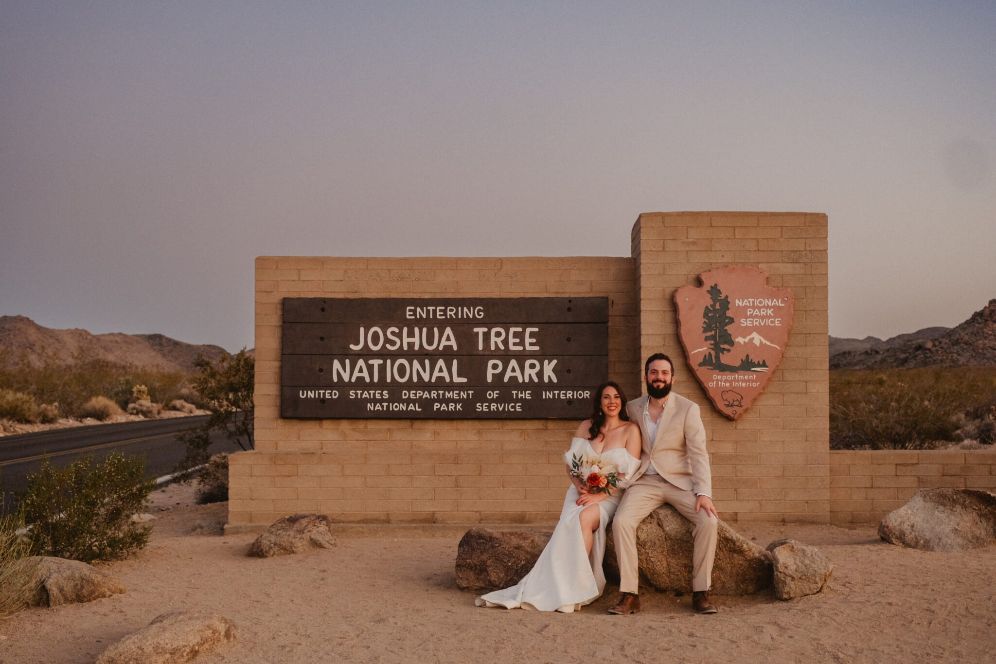 A couple dressed in wedding attire sits on a rock in front of the Joshua Tree National Park entrance sign at sunset.