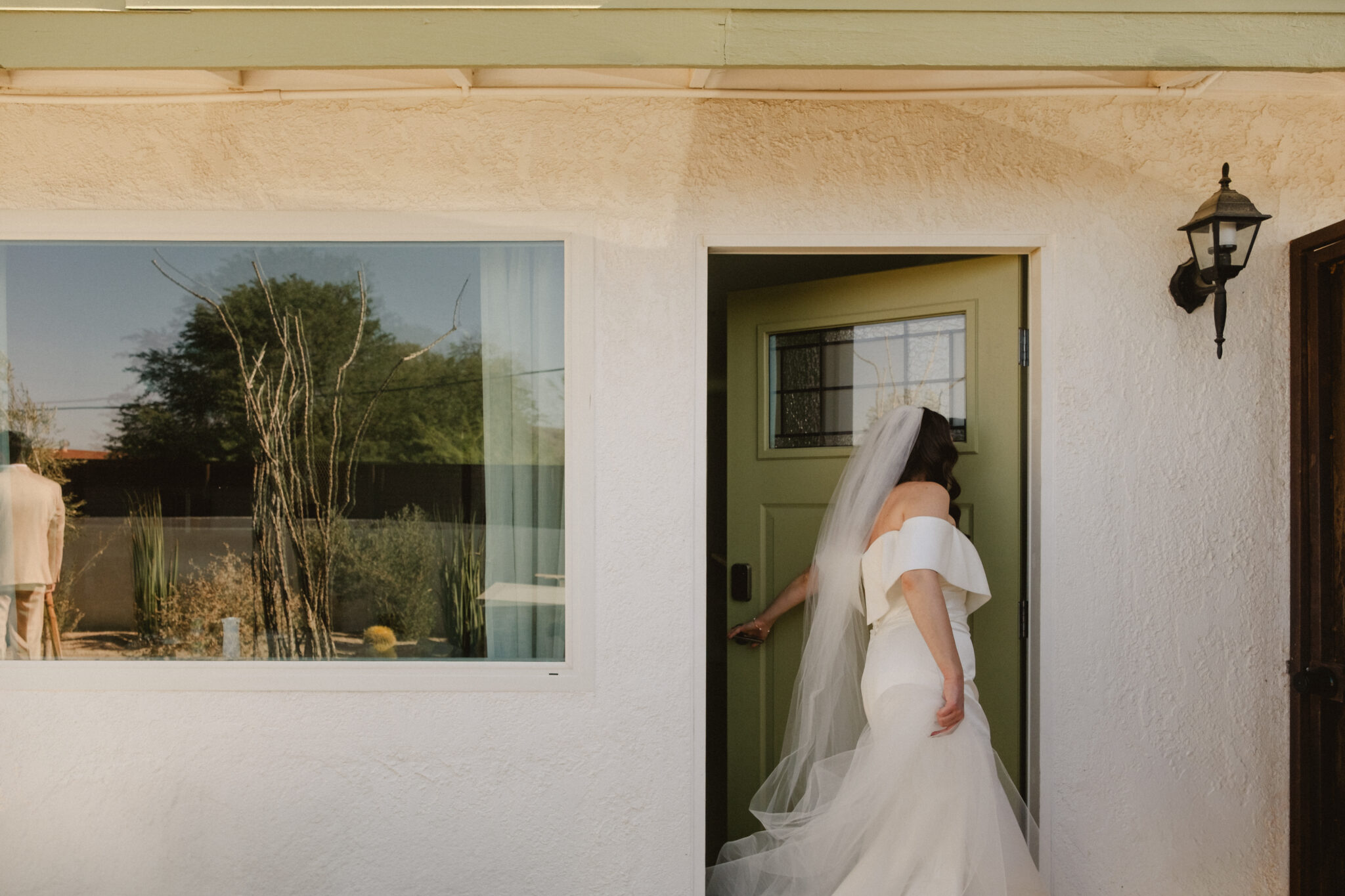 A bride in a white gown and veil enters a door of a white building with a lantern and a large window reflecting outdoor plants.