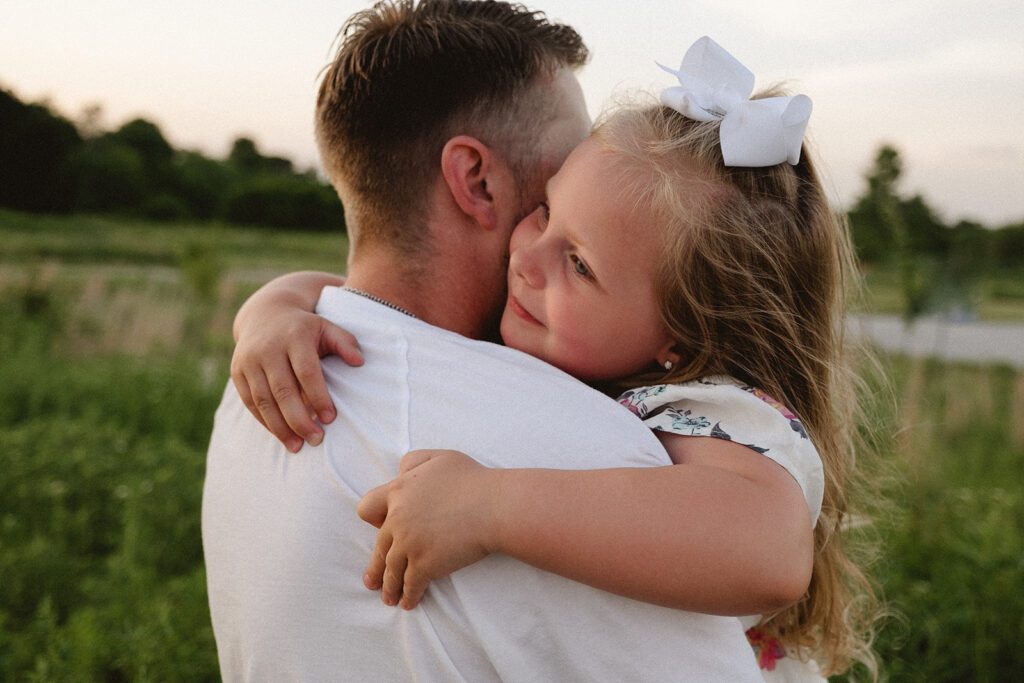 A young girl with a white bow hugs an adult man outdoors, both facing away from the camera in a grassy area.