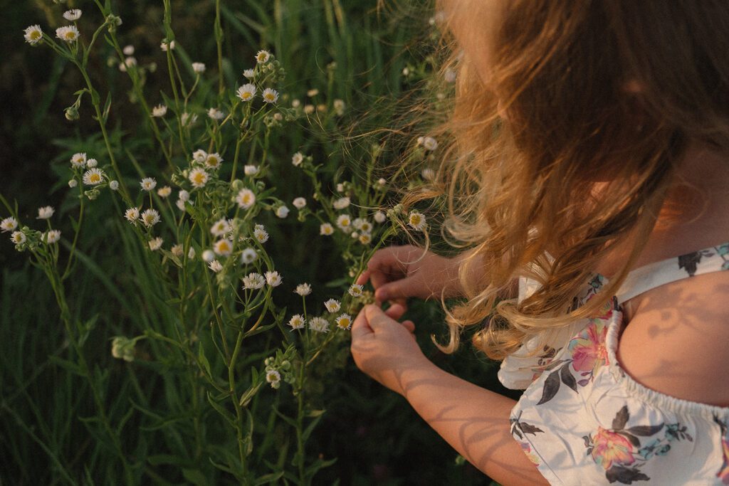 A child with wavy blond hair and a floral dress picks small white flowers from a green field in sunlight.