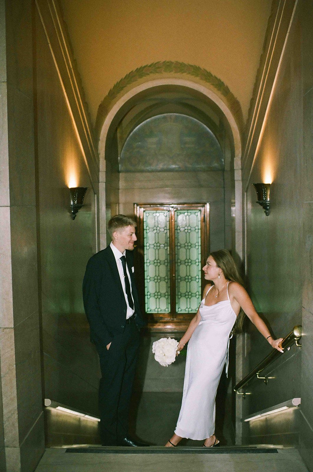 A man in a black suit and a woman in a white dress holding a bouquet stand on a marble staircase, facing each other, in an ornate, dimly lit hallway.
